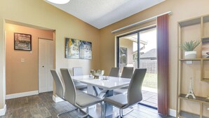 Un coin repas avec une table blanche et des chaises grises, un tableau au mur et une porte-fenêtre coulissante donnant sur un espace extérieur.