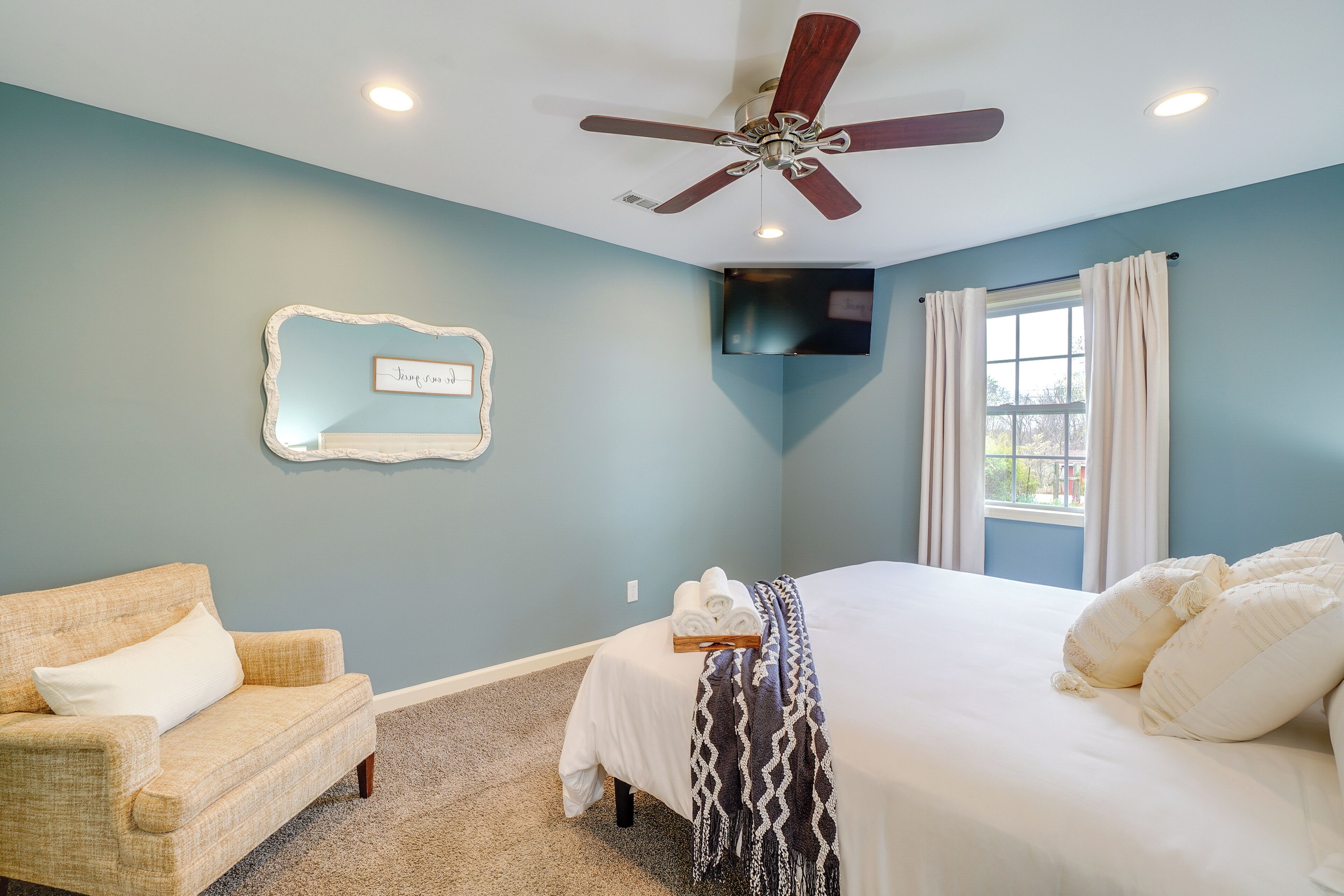 A bedroom with a bed, a chair, a ceiling fan, a TV, a window with curtains, and a wall-mounted mirror.
