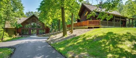 A log cabin with a porch and a deck, surrounded by trees and a paved driveway.