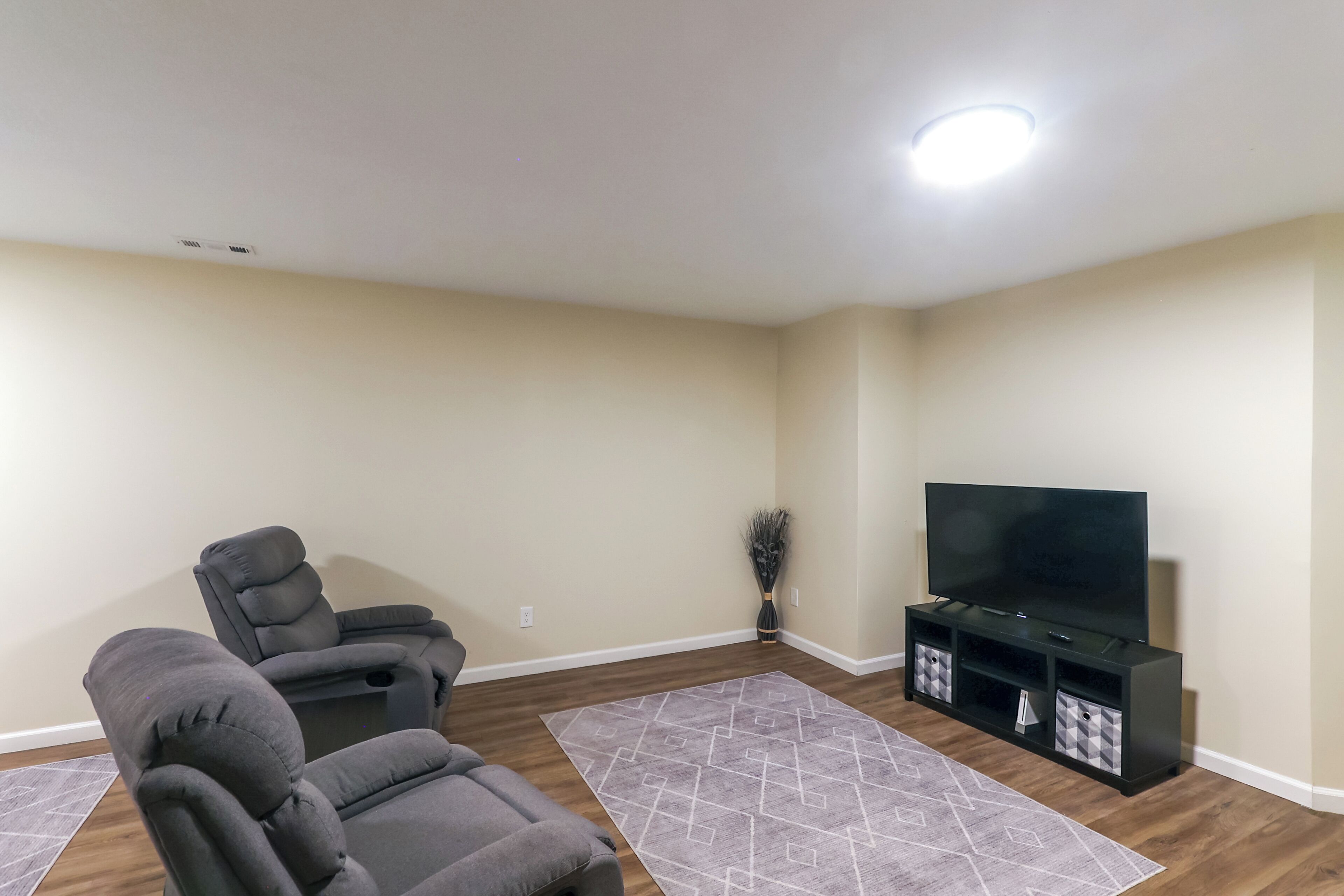 A living room with two gray recliner chairs, a flat-screen TV on a black stand, and a patterned rug on the wooden floor.