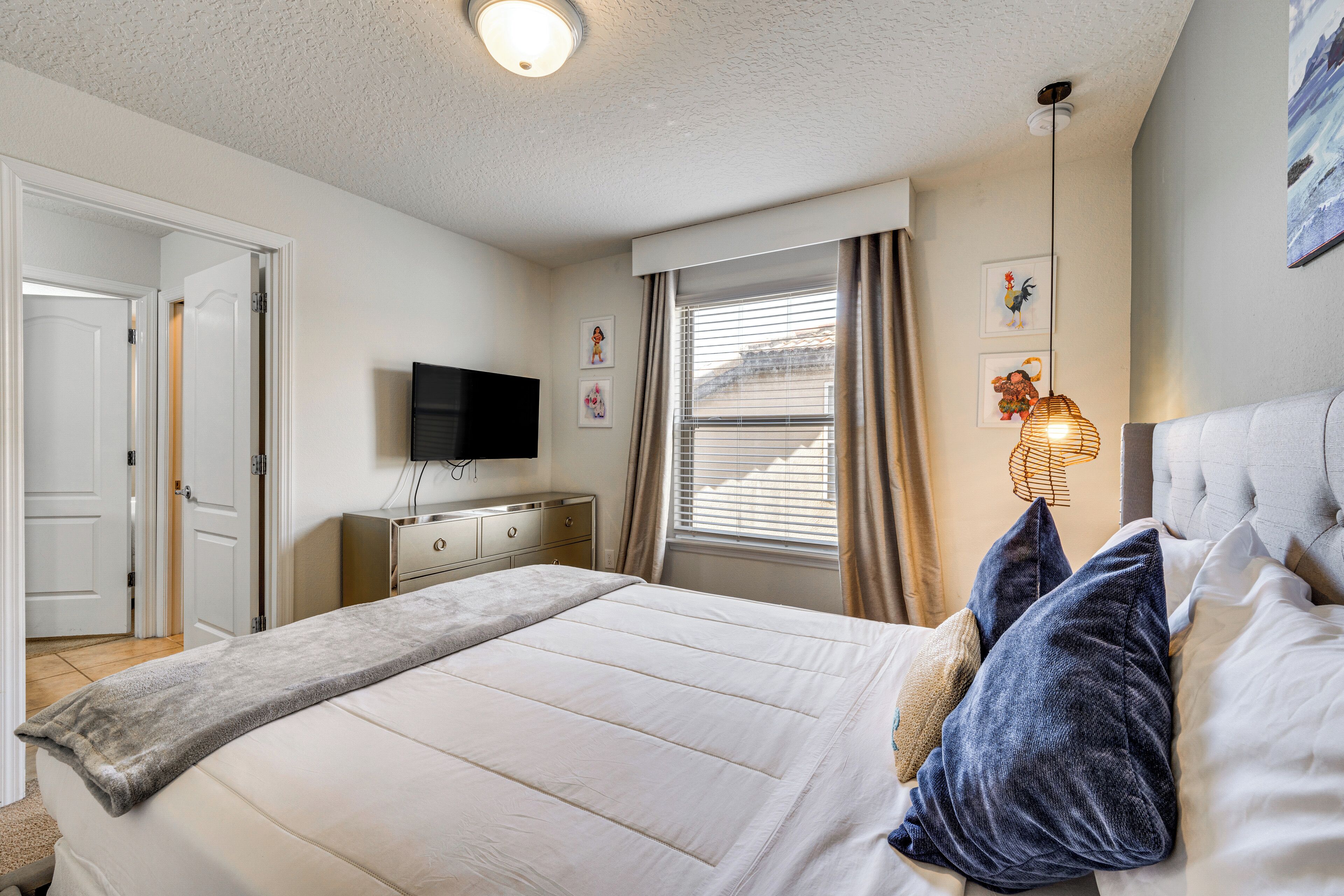 A neatly arranged bedroom with a bed, a television, a dresser, and a window with curtains.