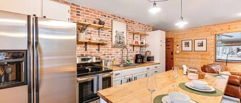Une cuisine moderne avec un réfrigérateur en acier inoxydable, un îlot central en bois surmonté d’une plaque de verre et un mur en briques.