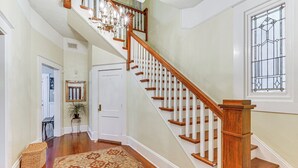 A staircase with wooden balusters and a chandelier, leading to a landing with a patterned rug and a potted plant.
