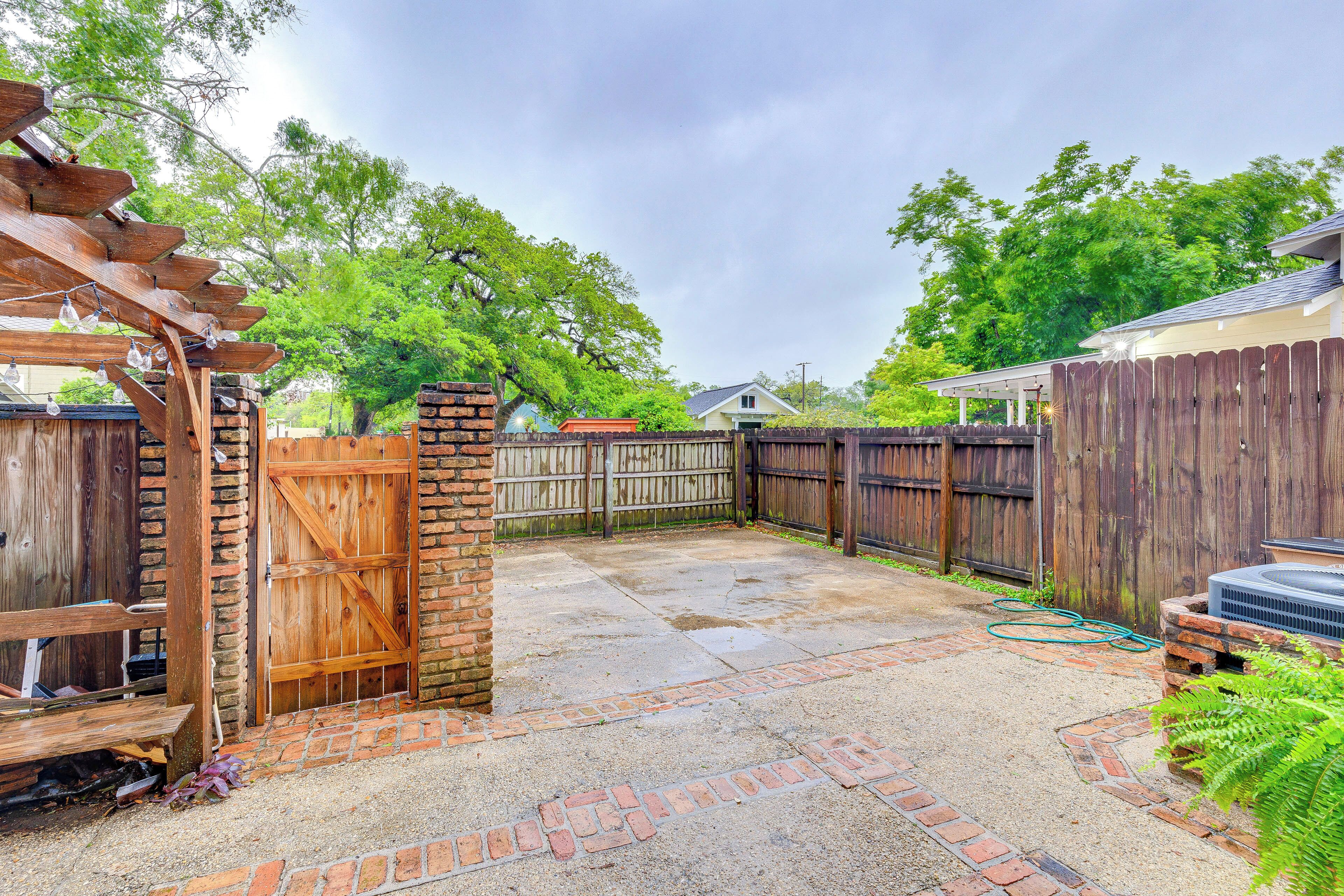 A backyard with a wooden pergola, a brick patio, a wooden fence, and a house in the background.