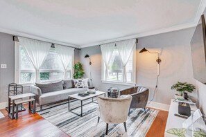Ett modernt vardagsrum med en grå soffa, ett soffbord och en grå fåtölj. Det finns två fönster med vita gardiner och en golvlampa.