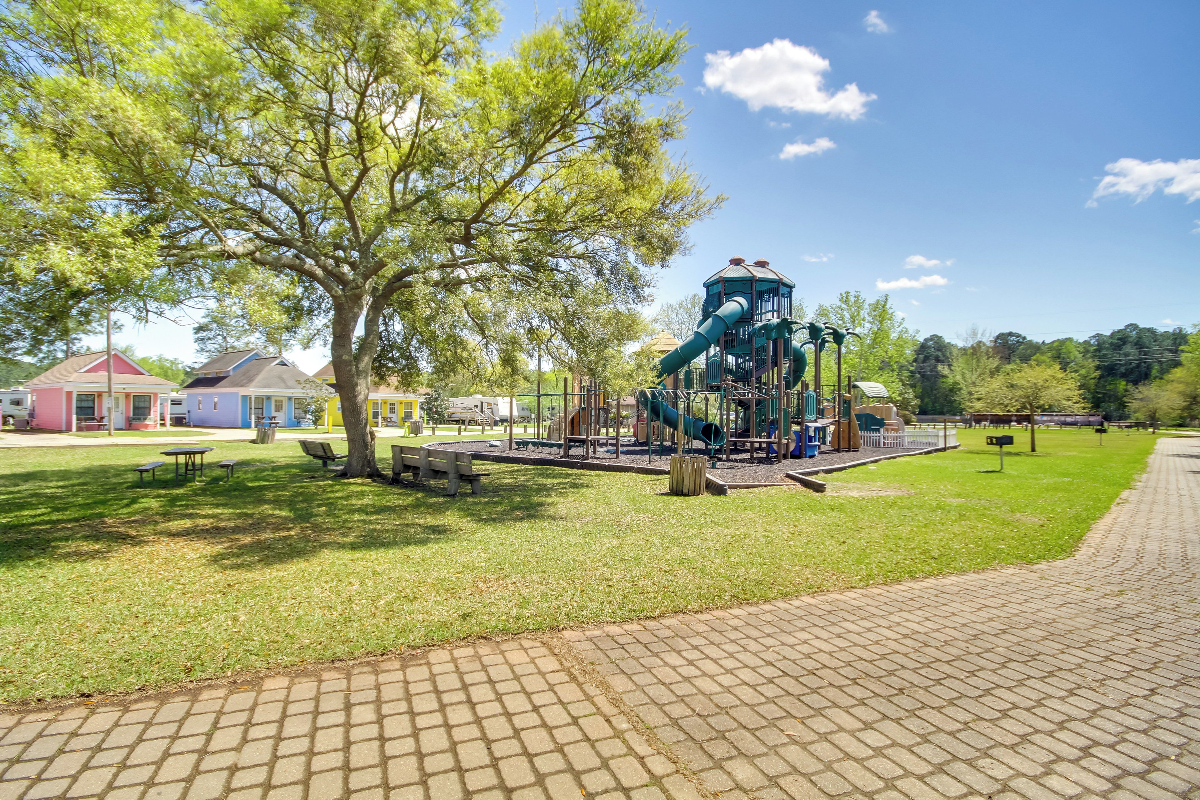 A playground with a slide and climbing structures, surrounded by grass and trees.