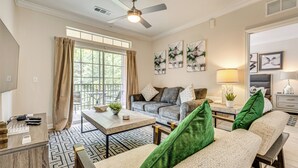 Ein helles Wohnzimmer mit einer Couch, einem Couchtisch und einem Deckenventilator. An der Wand hängen gerahmte Bilder und es gibt ein Fenster mit Vorhängen.