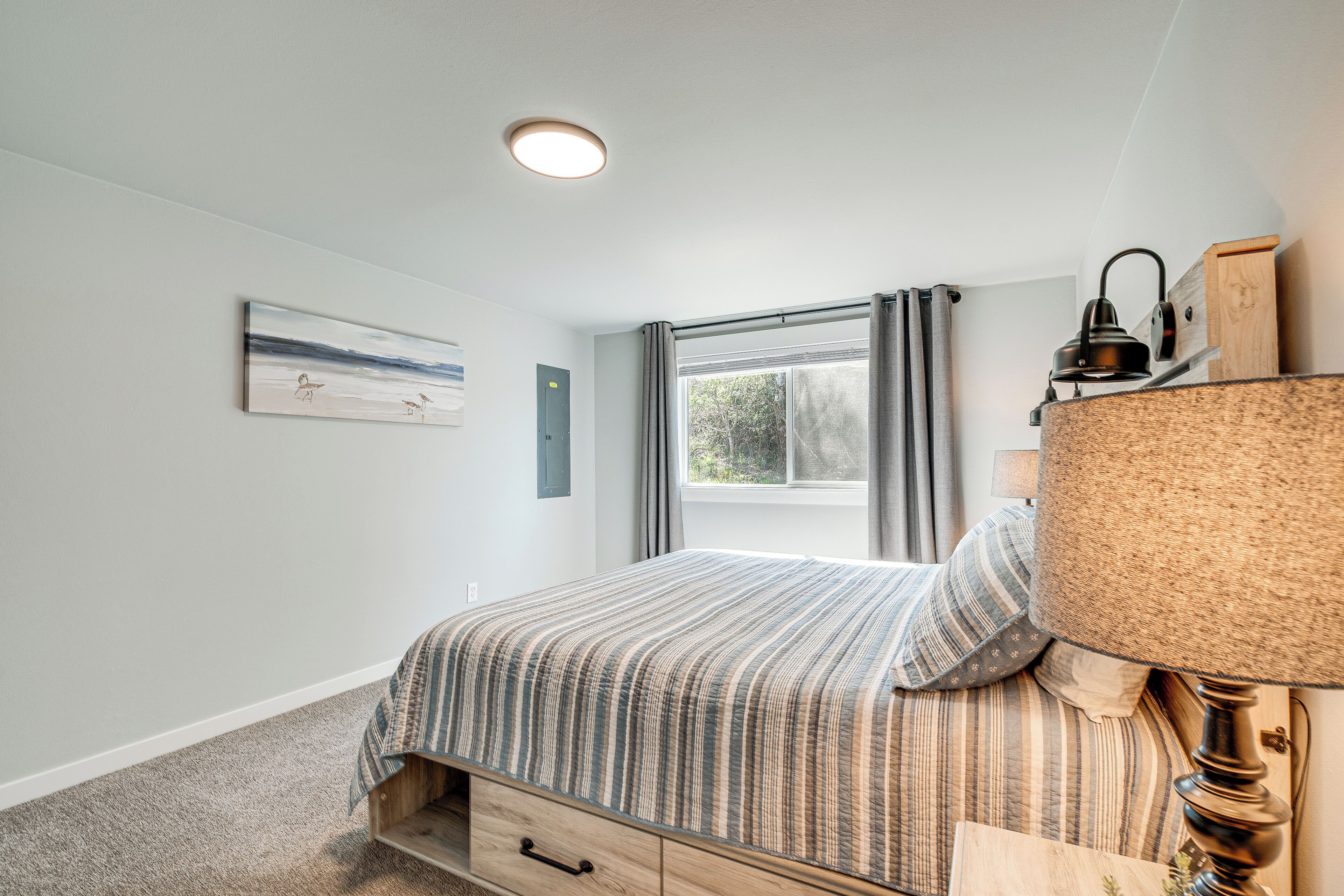 Une chambre à coucher avec un lit, une lampe, une fenêtre avec des rideaux, un tableau au mur et un luminaire au plafond.