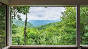 A view from a window with a wooden railing, overlooking a dense forest and distant mountains. - Sky Valley Retreat w/ Fire Pit & Mountain Views! (Dillard)