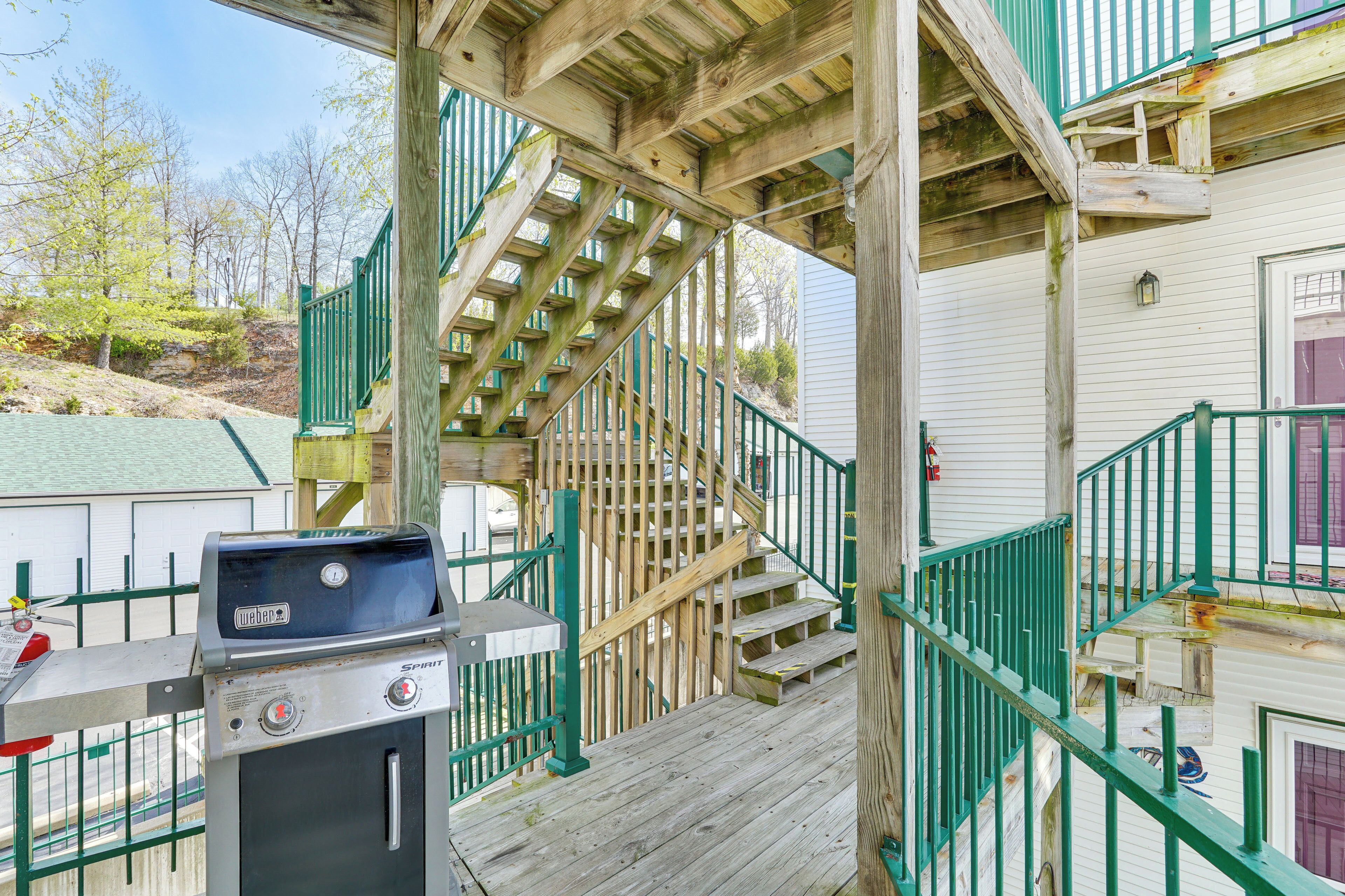 A wooden deck with a grill, a staircase with green railings, and a view of a building with a door and windows.