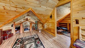 Une cabane en rondins avec un plafond en pente, des murs en bois, une cheminée centrale et un tapis à motifs sur le sol.