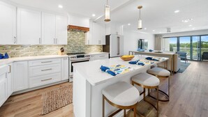 A modern kitchen with white cabinets, a central island with bar stools, a stone backsplash, and a large window offering a view of the outdoors. - 30a Hideaway 2 Bedroom Townhouse by RedAwning (Panama City Beach)