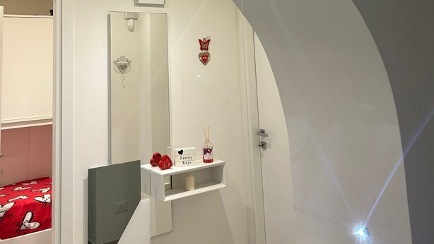 A bedroom with a curved ceiling, a bed with a red heart-patterned bedspread, a bedside table with a red vase, and a small shelf with a red candle and a sign that says "Family Keys". - Vacation House at Mely's (Marsala)