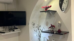A modern living room with a curved wall-mounted shelf holding decorative items, a wall clock, and a flat-screen TV on a stand. - Vacation House at Mely's (Marsala)