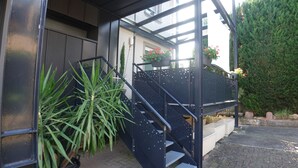 Un escalier menant à l’entrée d’un bâtiment, avec une rambarde en métal, des plantes en pot et un arbuste en arrière-plan.