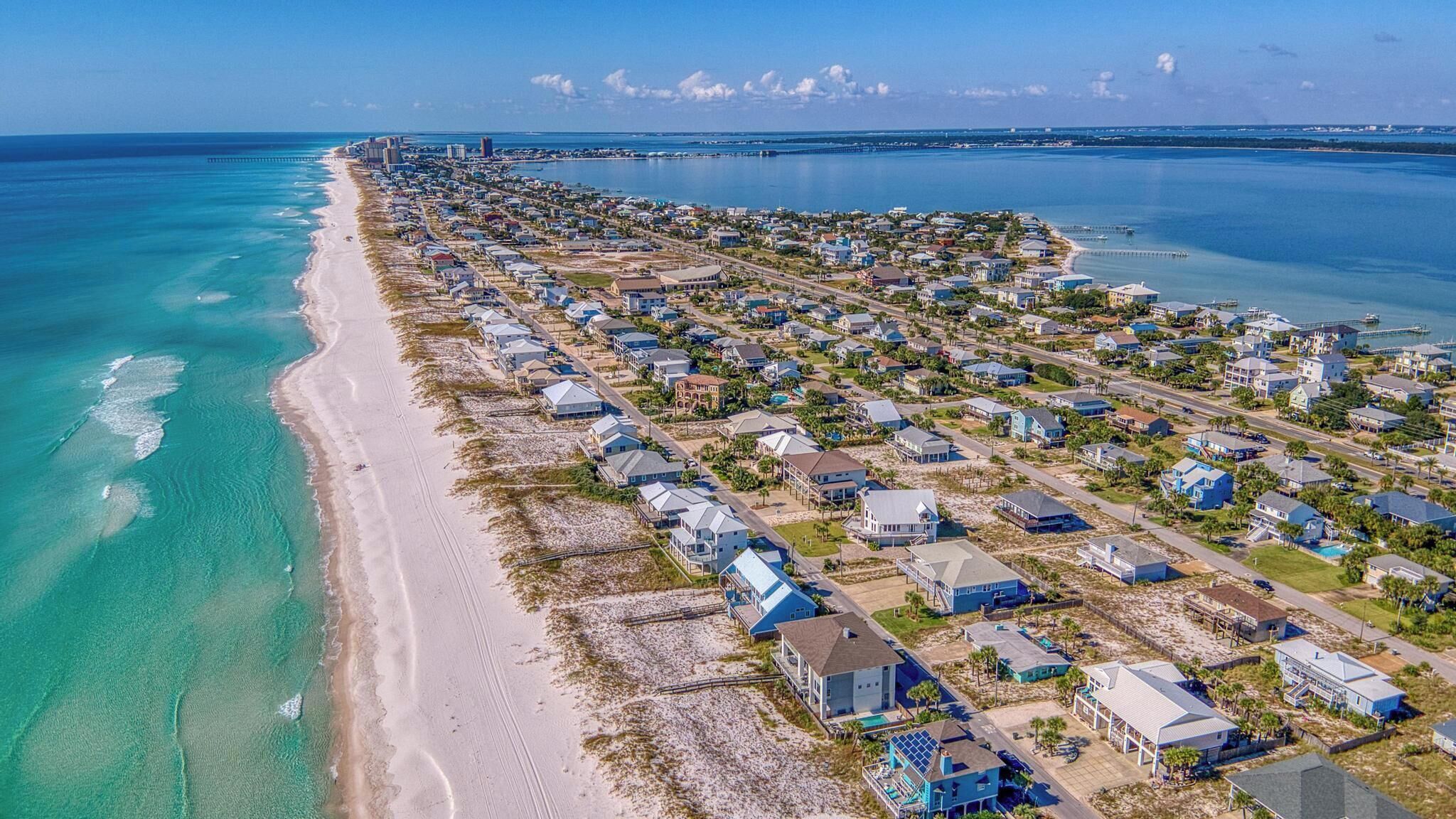Vista aérea de una zona residencial costera con casas a lo largo de la playa, aguas azules cristalinas y un muelle distante.