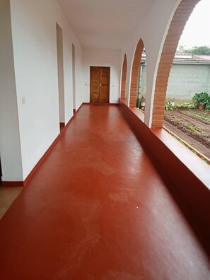 A long, red-tiled corridor with white walls and brick arches, leading to a wooden door. - Stc.Safari lodge (Karatu)
