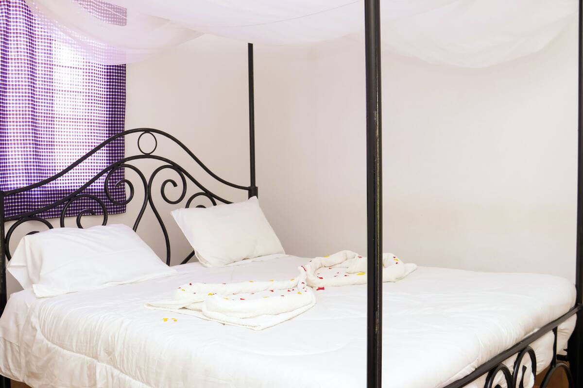 A four-poster bed with white linens and pillows, a checkered curtain, and a plain wall.