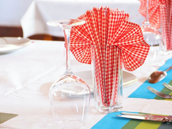 Una mesa con una copa de vino transparente, un vaso alto con servilleta de cuadros rojos y blancos y una cuchara sobre un mantel individual azul.