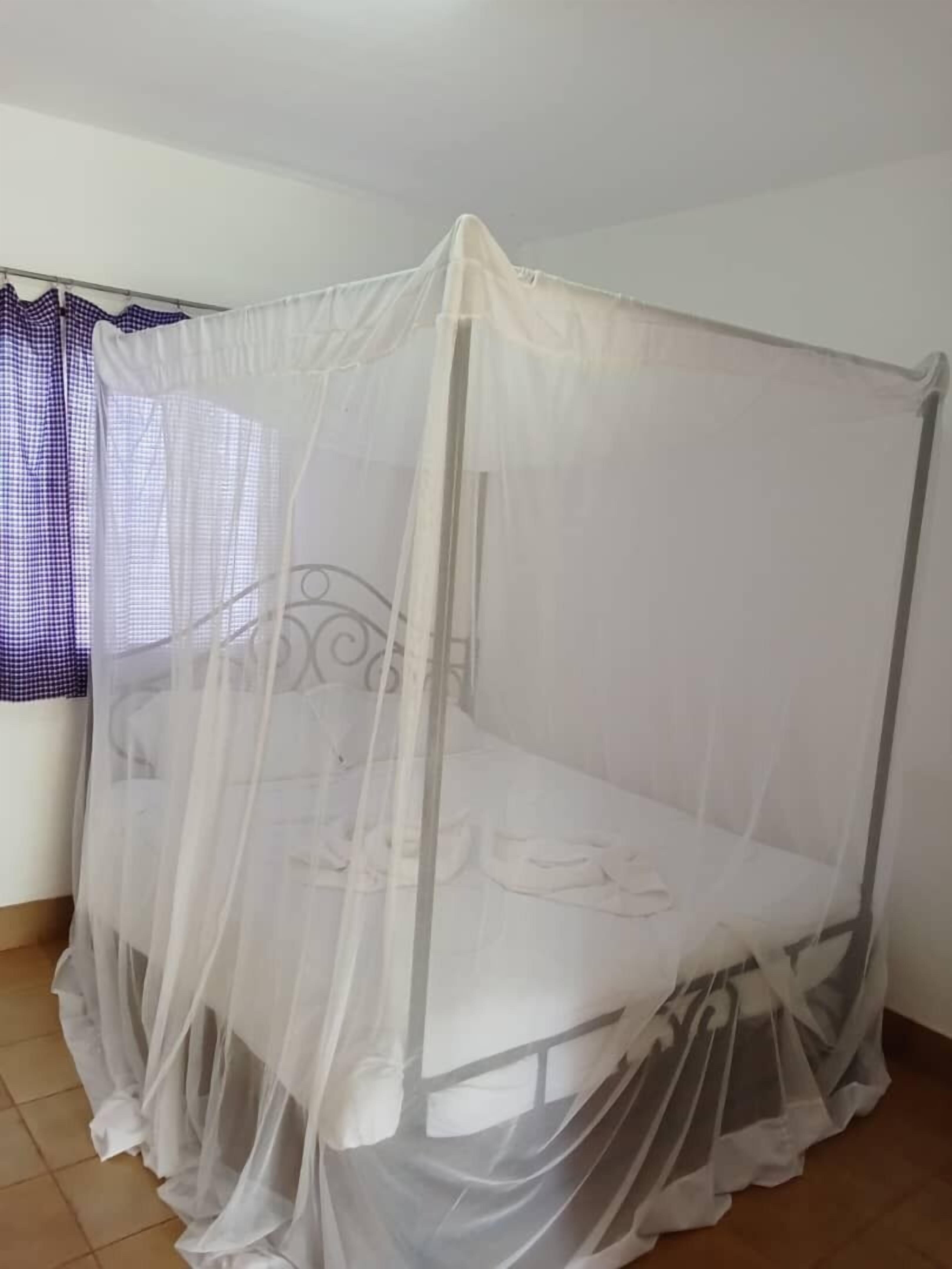 A single bed with a mosquito net in a room with white walls and a tiled floor.
