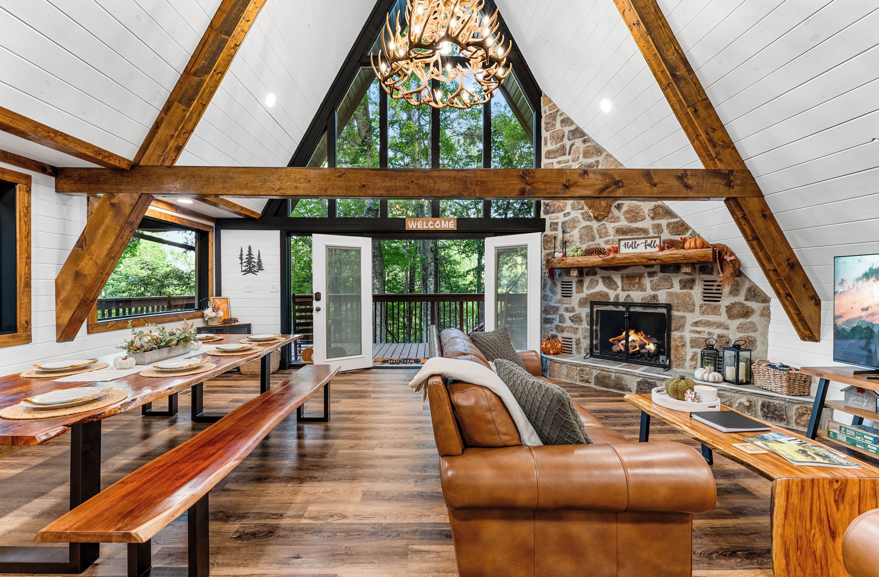 A spacious loft-style interior with a high ceiling, exposed wooden beams, a stone fireplace, and a large window overlooking greenery.