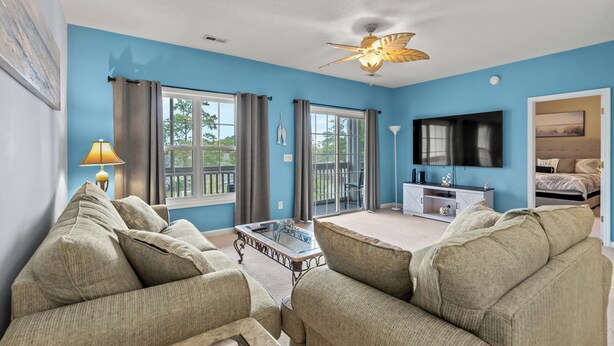 A living room with a blue wall, a ceiling fan, a flat-screen TV, and a sofa. - Short Stays in Summer - Pool - Seven Oaks 9-302 (North Myrtle Beach)
