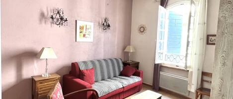 A living room with a sofa, chairs, a small table, a ceiling fan, a window with curtains, and a wall with a chandelier and pictures.