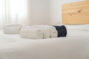 Ein ordentlich bezogenes Bett mit aufgerollten weißen und dunkelblauen Handtüchern, einem hölzernen Kopfteil und einer weißen, strukturierten Bettdecke.