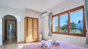 A bedroom with a bed, a wardrobe, an air conditioning unit, and a window with a view of greenery.