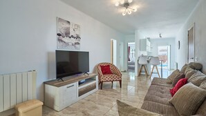 A living room with a TV, a sofa, a chair, and a dining area in the background.