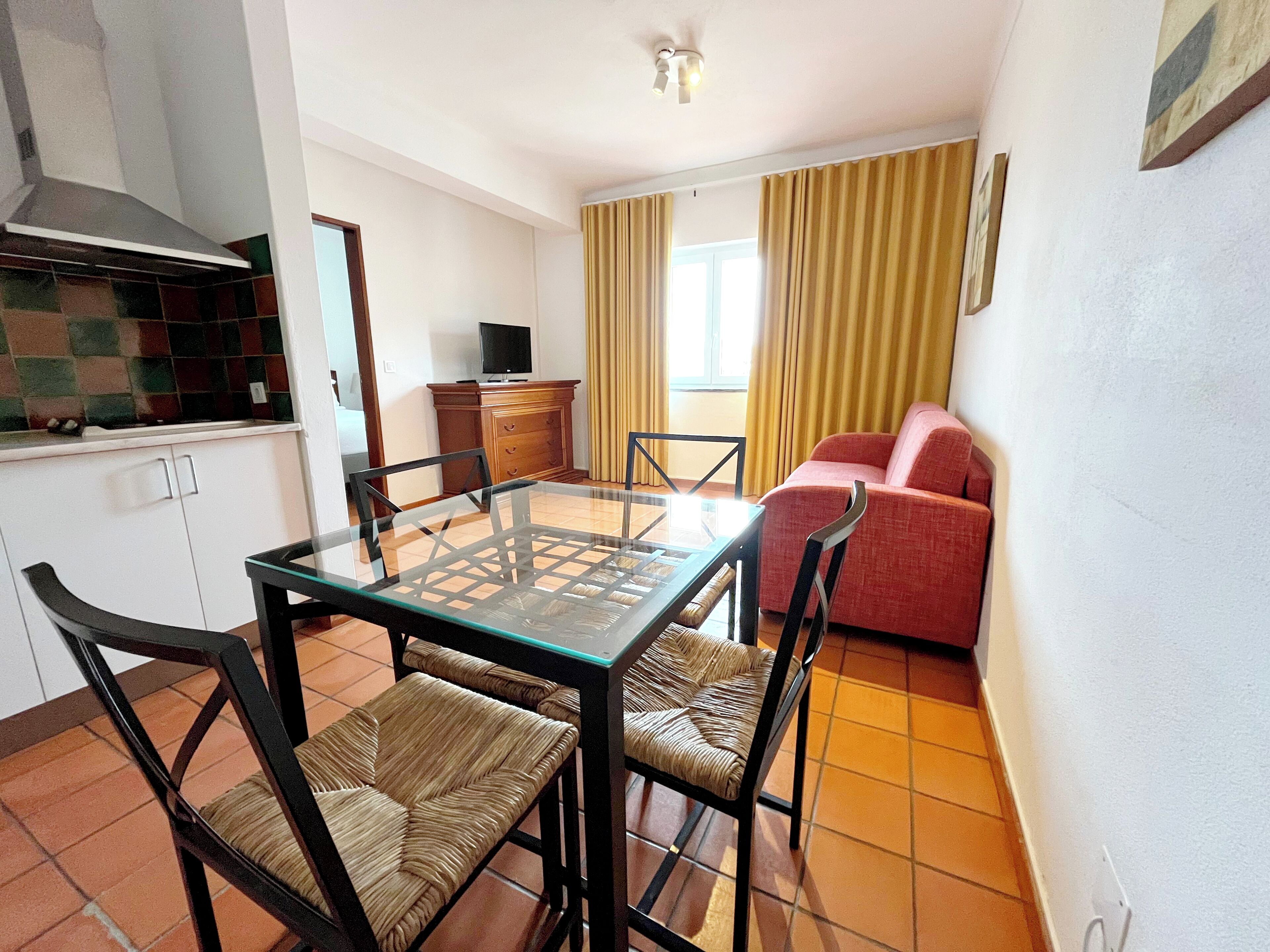 Un espacio habitable compacto con una mesa de comedor, sillas, un sofá, un televisor sobre un mueble y una zona de cocina.