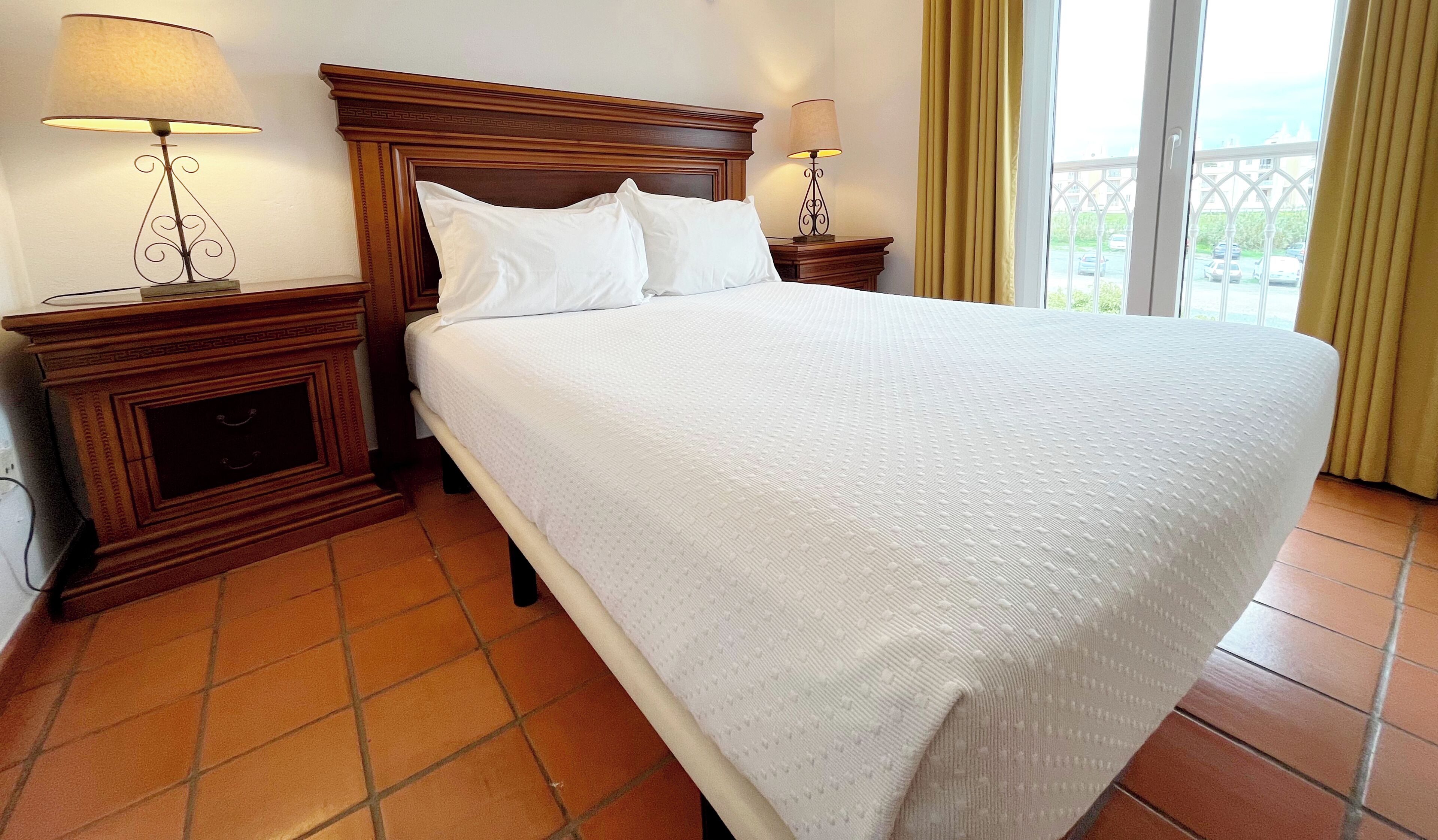 Una cama bien hecha con sábanas blancas, dos mesitas de noche con lámparas y vistas a un balcón a través de una ventana con cortinas.