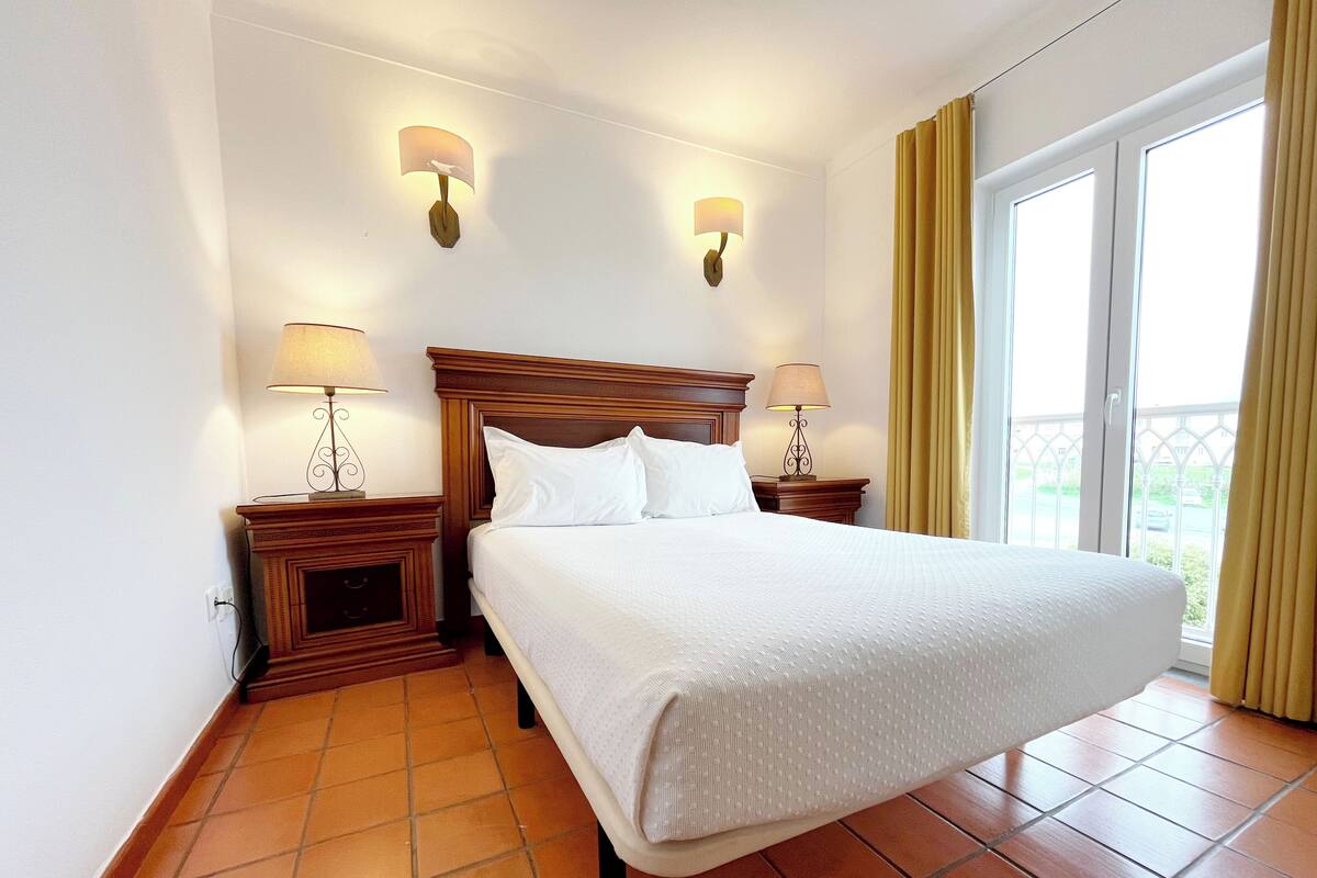 A neatly made bed in a hotel room with two bedside lamps, a wooden headboard, and a large window with curtains.