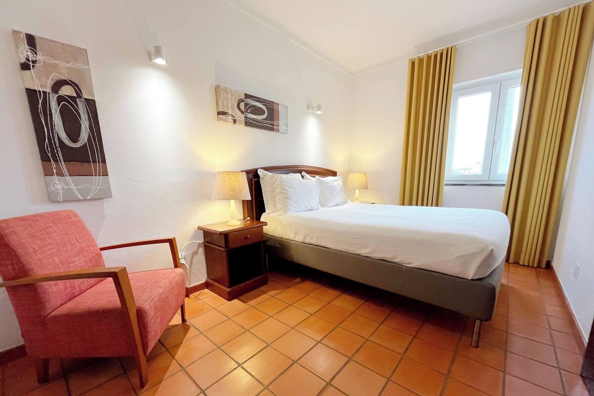 A hotel room with a bed, a chair, a nightstand with a lamp, and artwork on the walls.