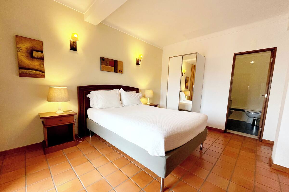 A hotel room with a large bed, two bedside tables with lamps, a mirror, and a door to the bathroom.