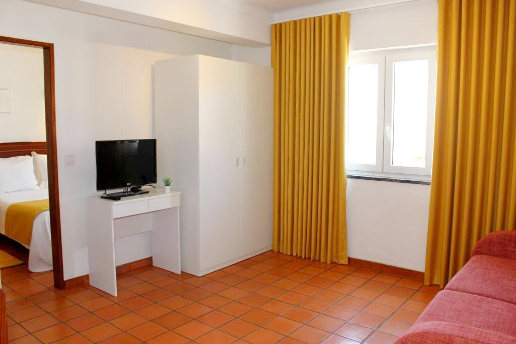 A hotel room with a bed, a desk with a TV, a wardrobe, and a window with yellow curtains.