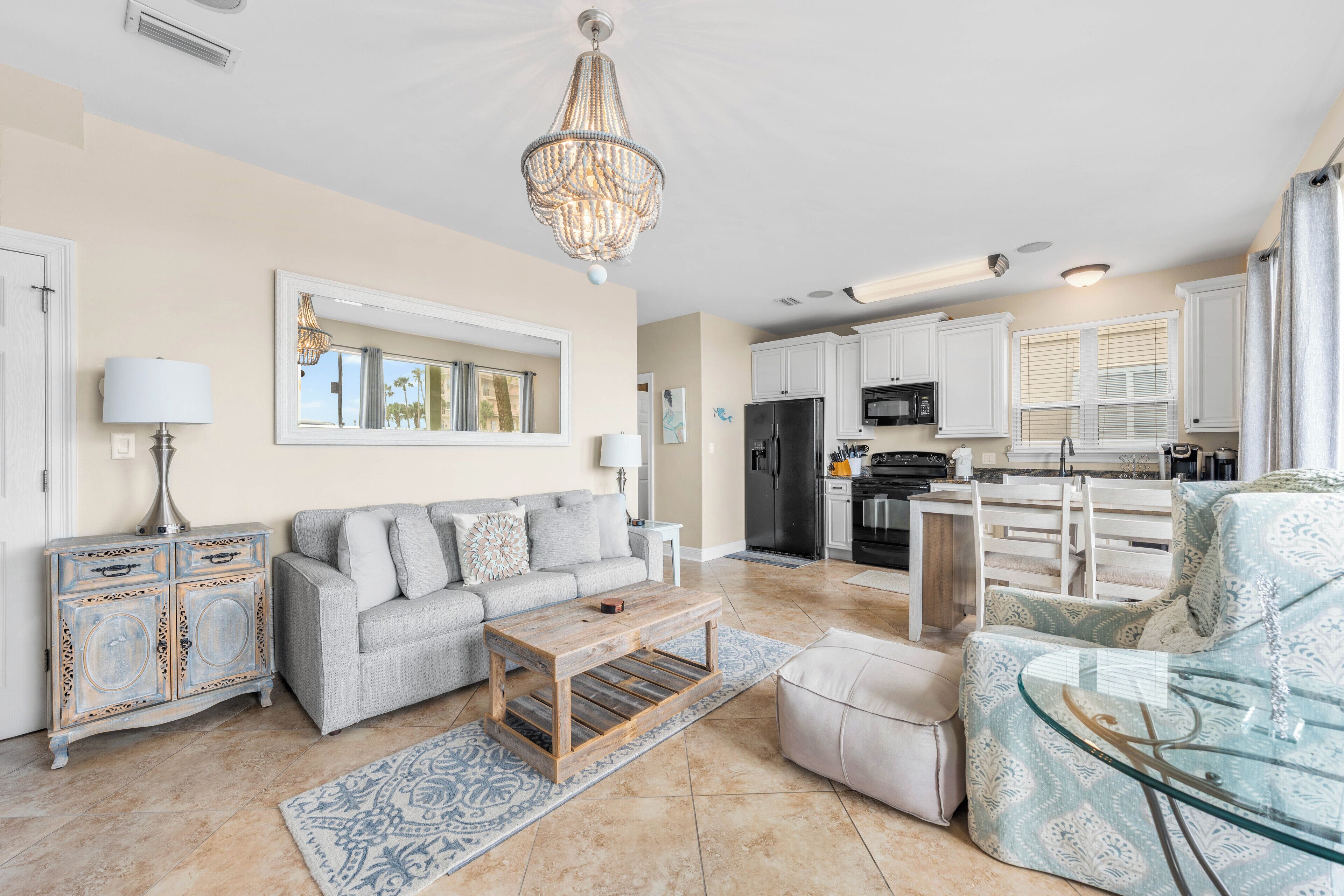 A spacious living room with a sofa, coffee table, and ottoman, a dining area with a glass table, and a kitchen with white cabinets and appliances.