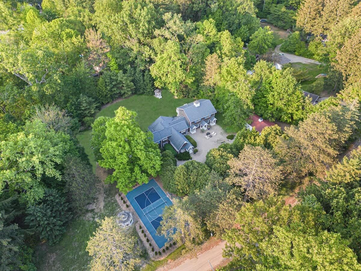 An aerial view of a house surrounded by dense forest, with a tennis court and a driveway leading to the entrance.