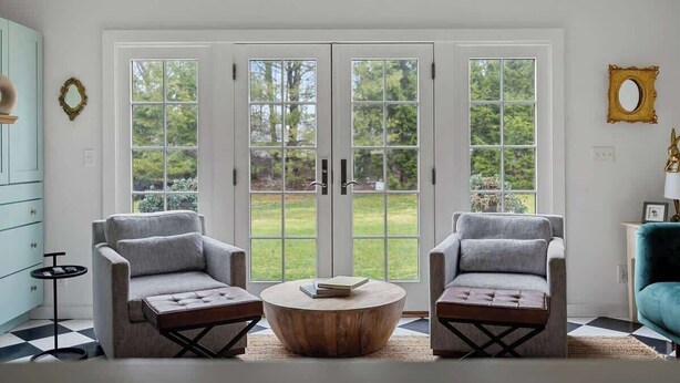Una habitación con dos sillones grises, una mesa de centro redonda de madera y vistas a un jardín a través de amplios ventanales.