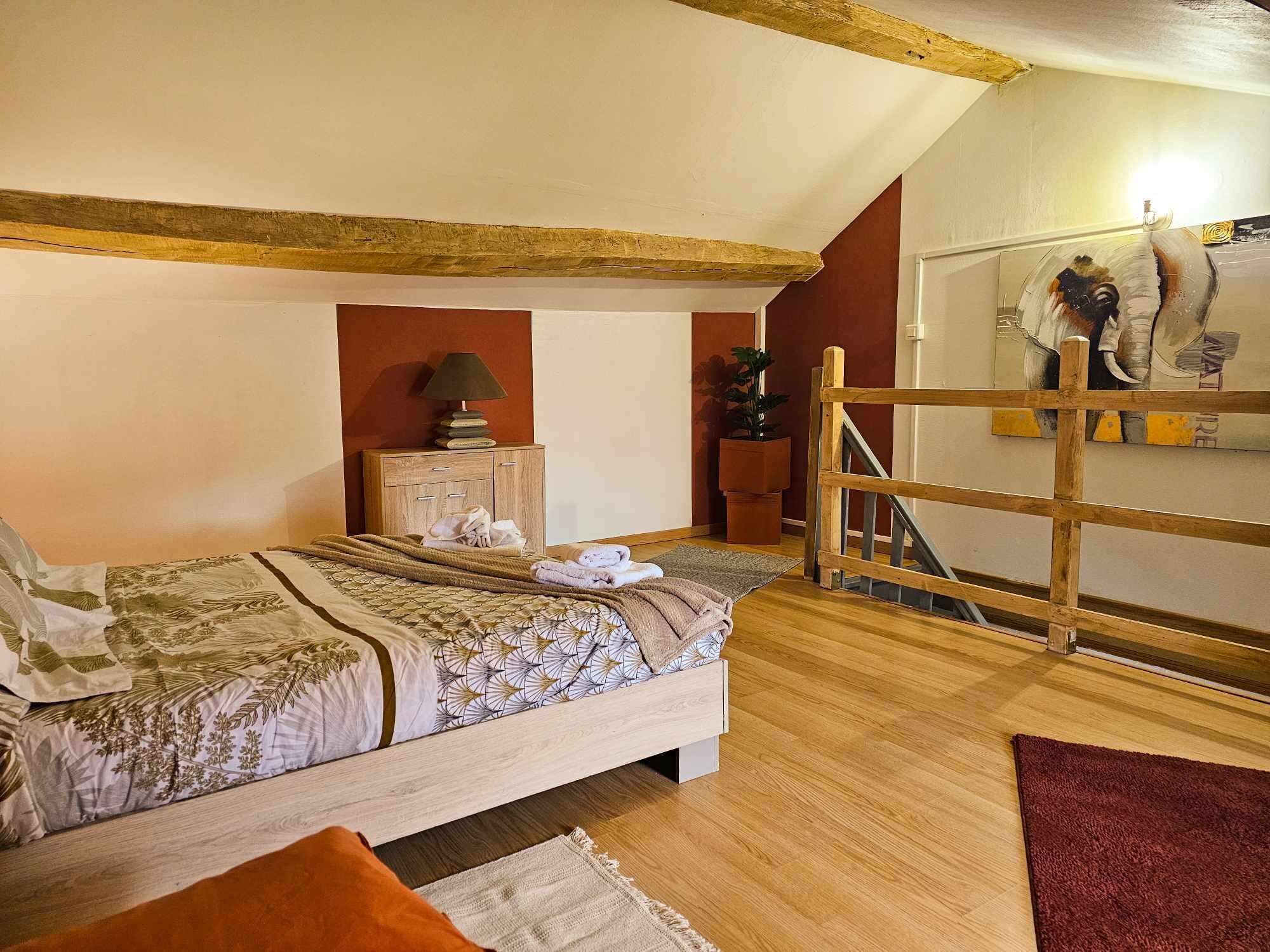 Un dormitorio con una cama de madera, una mesita de noche con una lámpara, una pequeña mesa con libros y una planta en maceta.