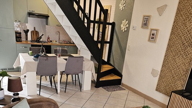 A modern living room with a staircase, a dining area, and a kitchen in the background. - Maison en Centre-ville Avec Jardin Privatif (Nevers)