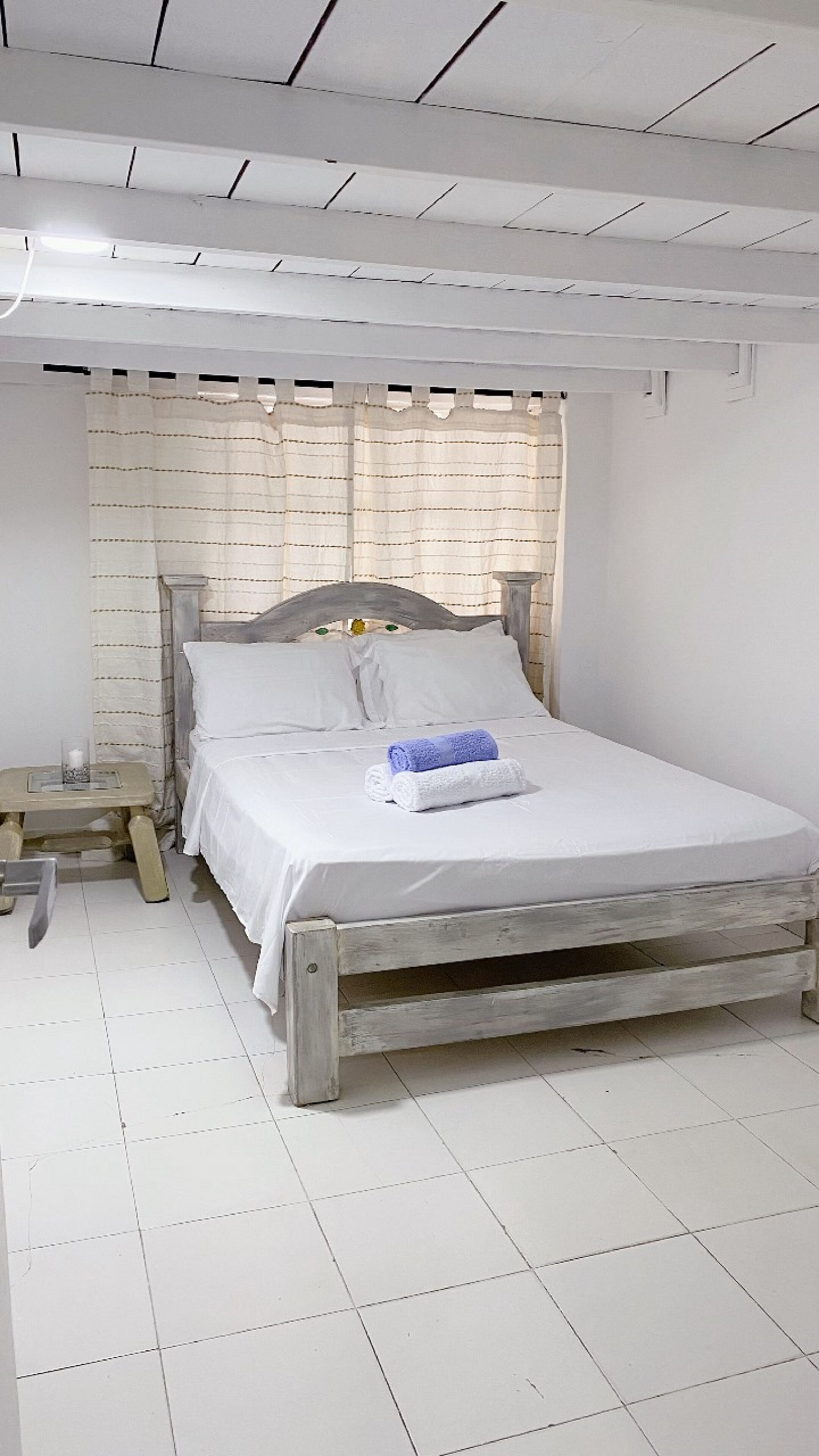 Un lit simple avec un cadre en bois et du linge de lit blanc, une petite table de chevet avec une lampe, et une fenêtre avec des rideaux à rayures.
