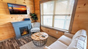 A cozy cabin interior with a fireplace, a sofa, a round coffee table, and a large window with blinds.