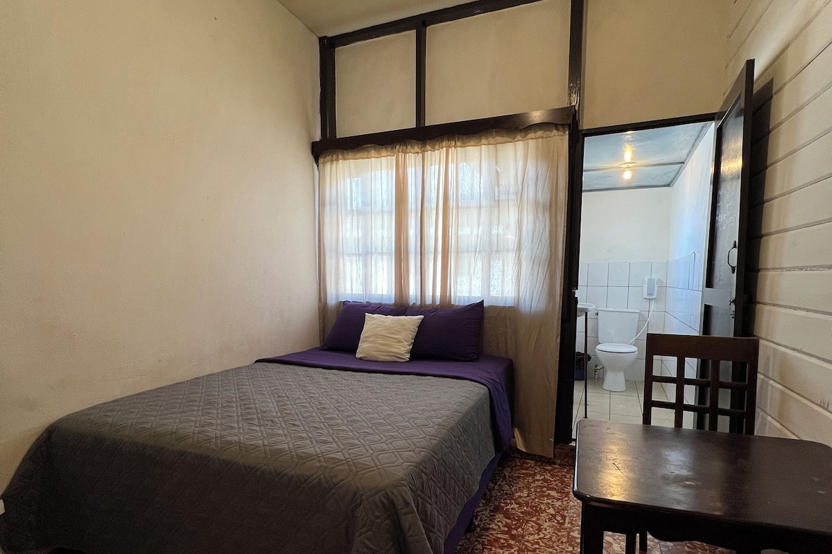 Une chambre à coucher avec un lit, une chaise, une table et une salle de bain visible à travers une porte.