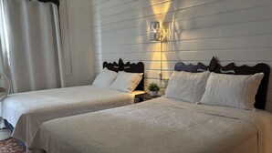 Dos camas matrimoniales con ropa blanca, un aplique de pared y una pequeña planta en maceta sobre una mesita de noche.
