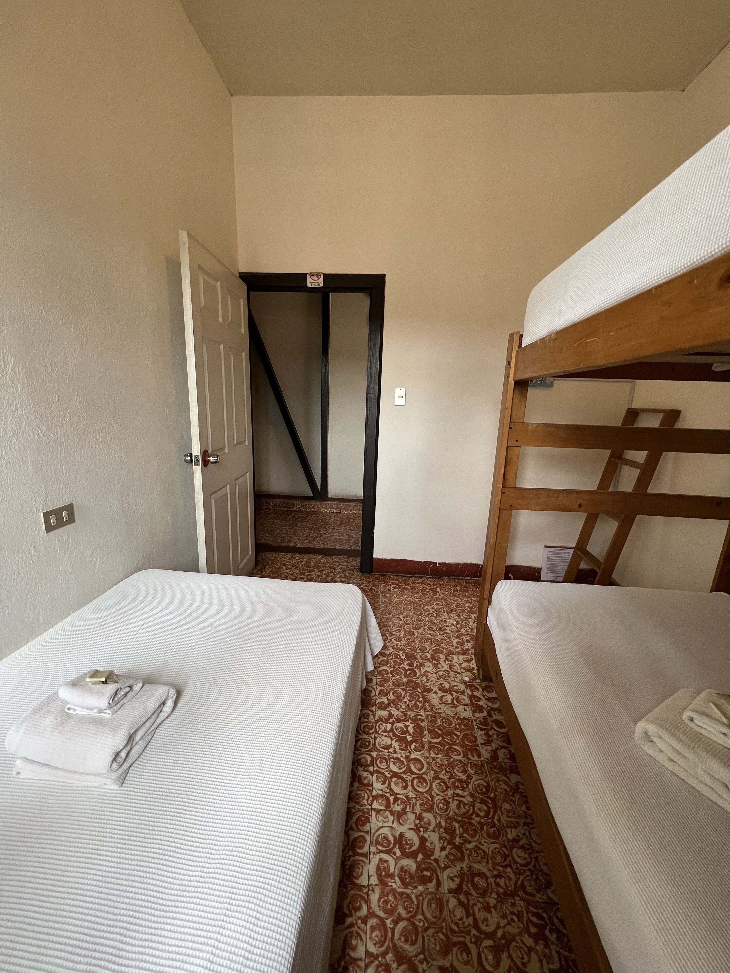 Una habitación con dos camas, una con estructura de madera y otra de metal, ambas con ropa blanca. Hay una puerta que conduce a otra habitación y una alfombra estampada en el suelo.