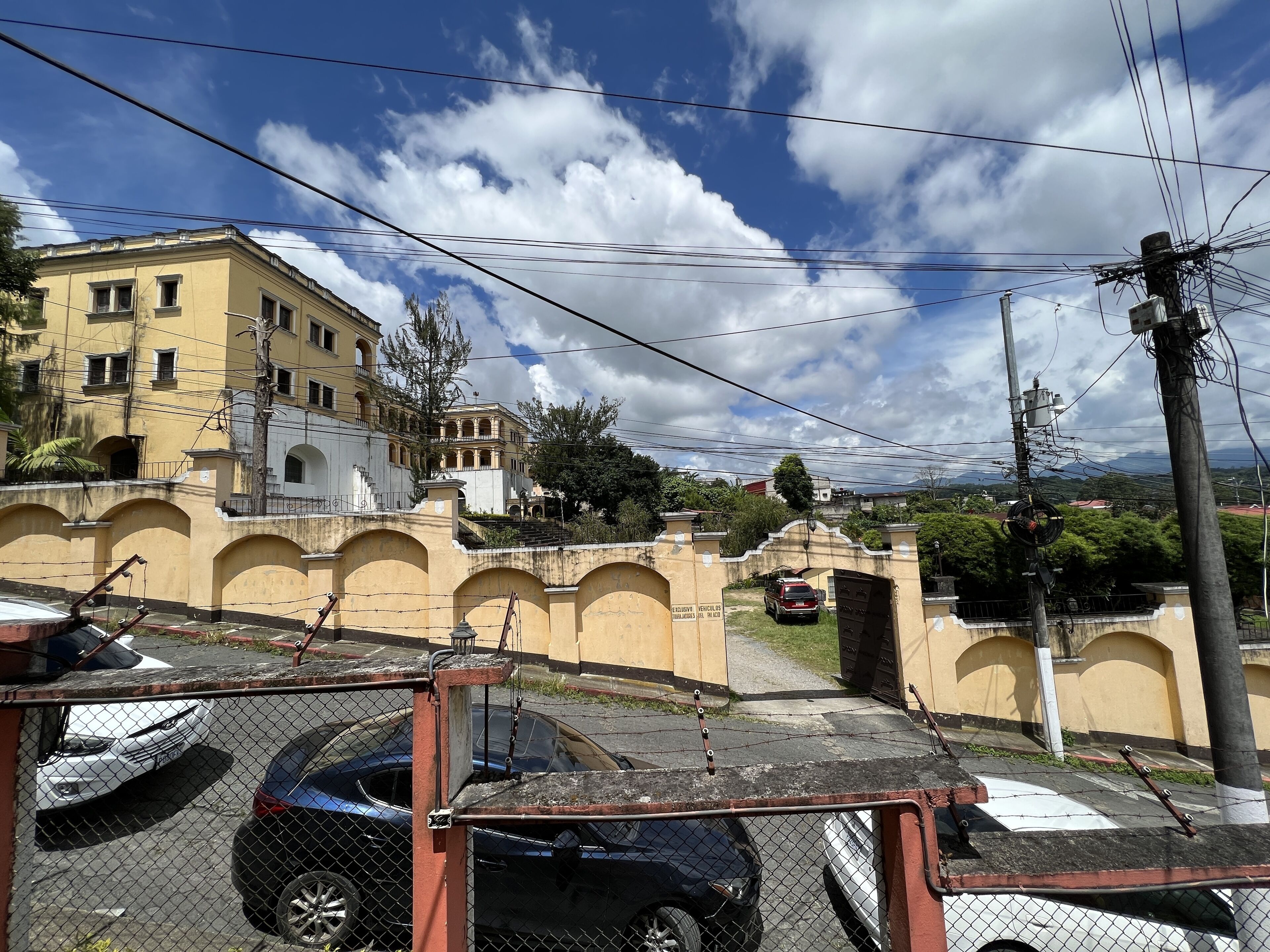 A fenced parking area with several cars, a yellow building with arched windows, and a mountainous landscape in the background.