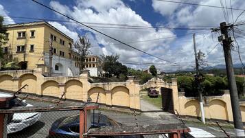 A fenced parking area with several cars, a yellow building with arched windows, and a mountainous landscape in the background.
