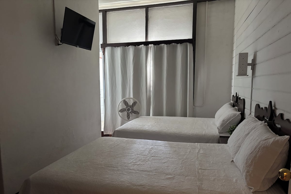 Une chambre avec deux lits, un téléviseur fixé au mur, une fenêtre avec des rideaux et un ventilateur fixé au mur.