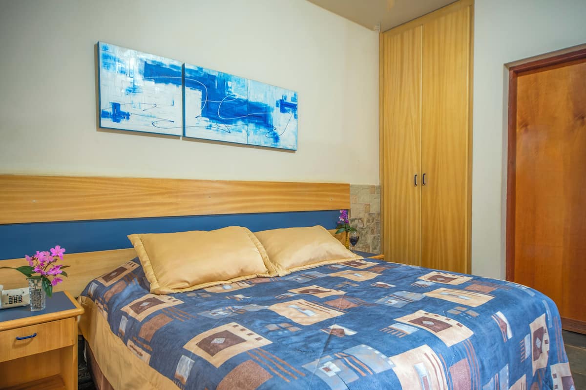 Un dormitori amb un capçal de llit de fusta, un edredó estampat, una tauleta de nit amb un telèfon i un ram, i un quadre abstracte blau a la paret.